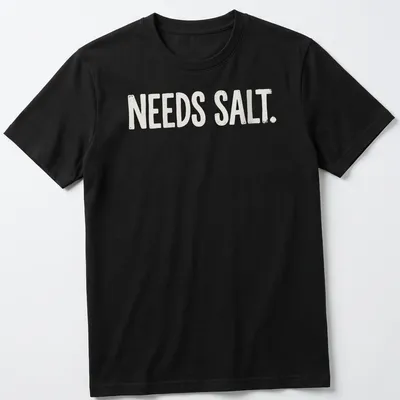 Bold distressed chef graphic with the text 'NEEDS SALT.' in white and light beige — printed on a unisex t-shirt