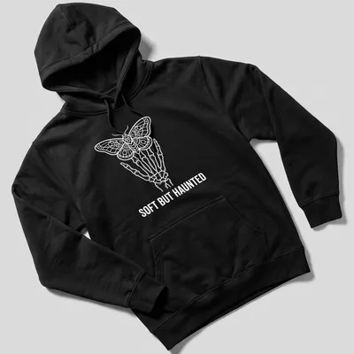 Line art of a skeleton hand holding a moth with the text 'SOFT BUT HAUNTED' in white. — printed on a unisex hoodie
