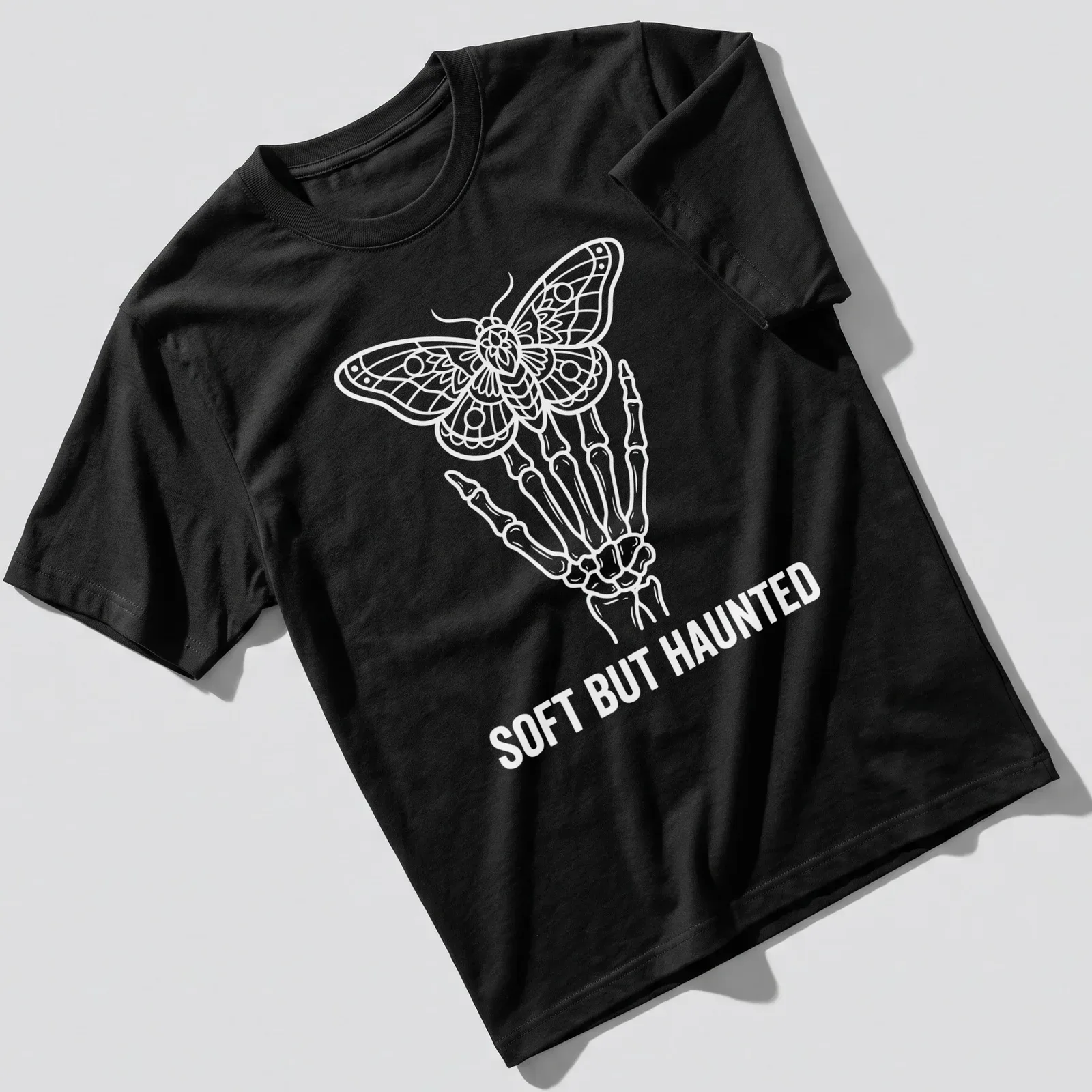 Line art of a skeleton hand holding a moth with the text 'SOFT BUT HAUNTED' in white. — printed on a unisex t-shirt