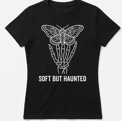 Line art of a skeleton hand holding a moth with the text 'SOFT BUT HAUNTED' in white. — printed on a women's shirt